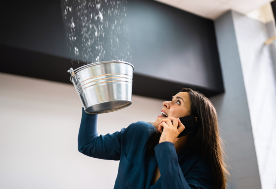 Woman catching water from a leaking ceiling in a bucket while calling for help on her phone.
